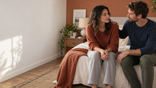 Two adults sit on a bed and talk, with an unopened condom packet on the bedside table.