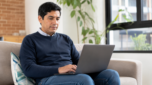 A middle-aged man wearing a dark blue sweater and jeans sits on a light-colored couch, focused on a dark grey laptop resting directly on his lap. The background features a bright room with a potted plant, a brick wall, and a window.
