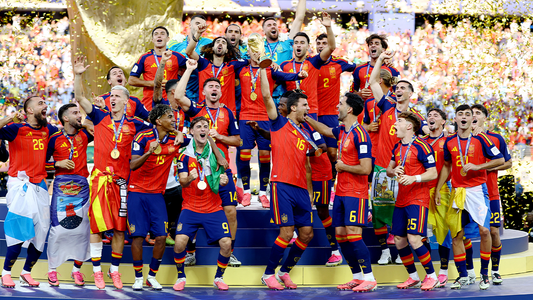 The Spain national football team, as featured in worldcup.jpg, celebrates winning the World Cup trophy on a podium. Players in red and blue kits are cheering, with one raising the trophy, while golden confetti falls in a crowded stadium.